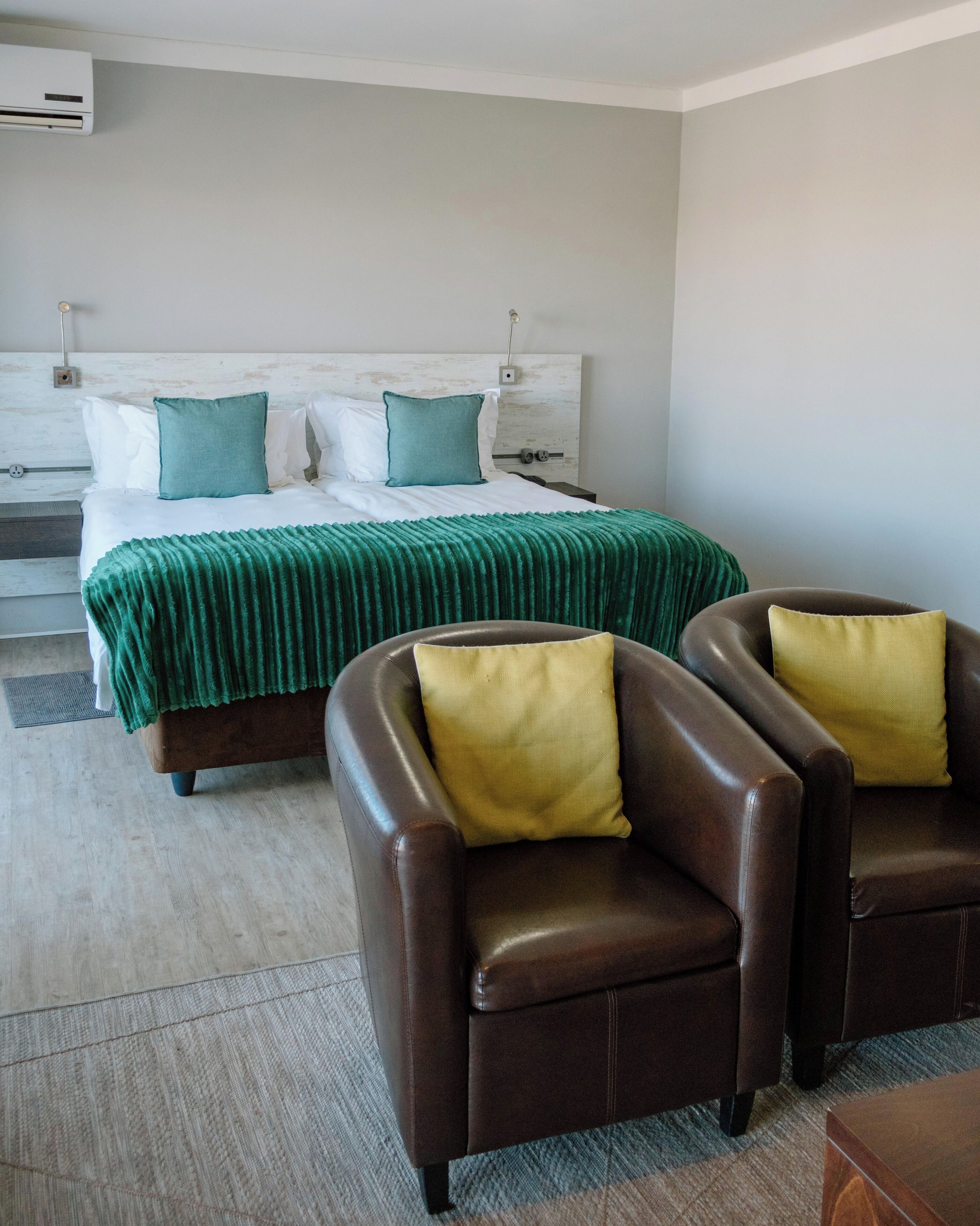 Een hotelkamer met een bed, twee fauteuils, een nachtkastje en een wandairconditioner.