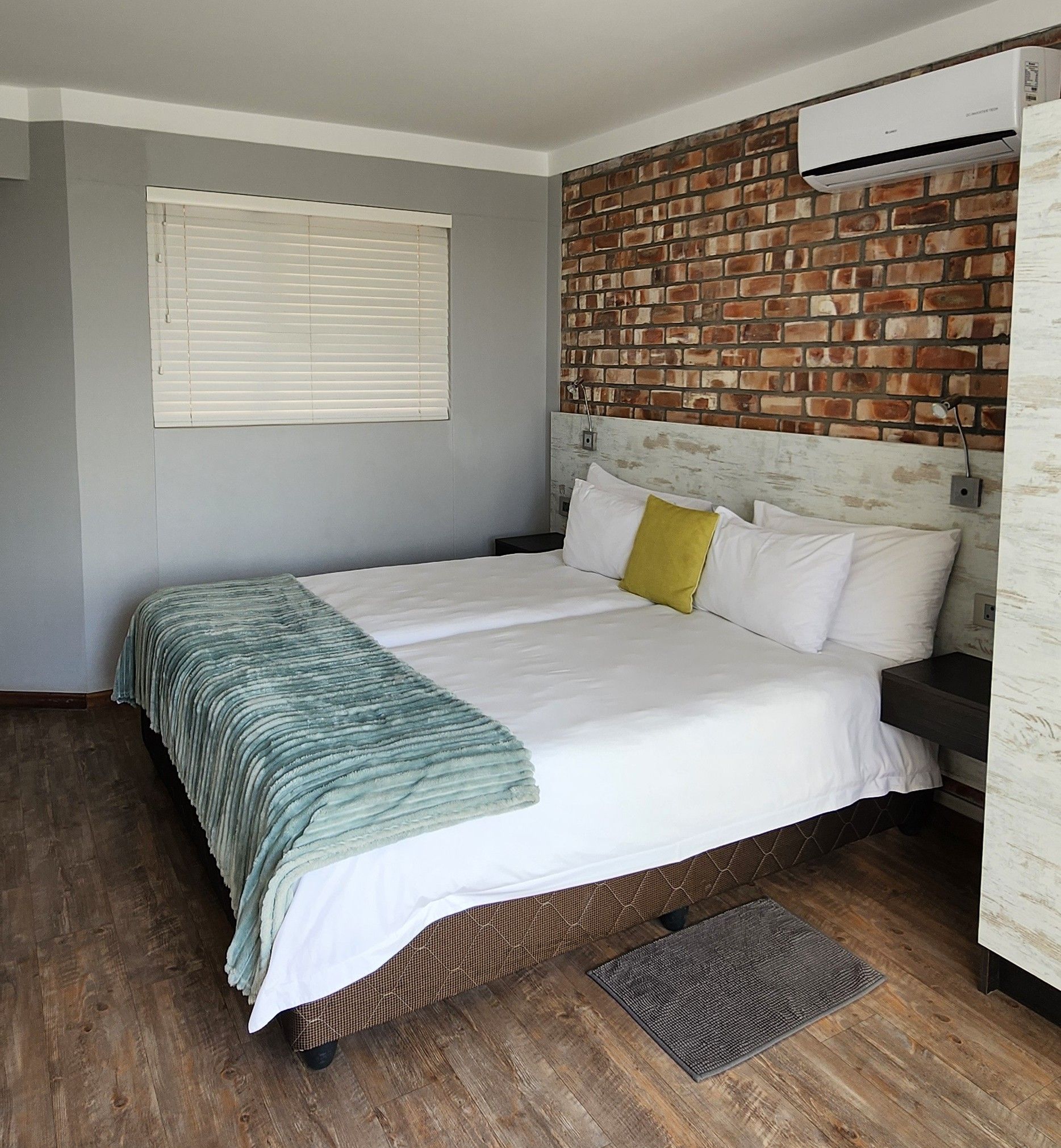 A hotel room with a large bed, a brick-patterned headboard, a window with blinds, and a wall-mounted air conditioning unit.
