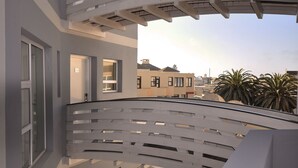 A modern balcony with a white railing, a glass door, and a view of neighboring buildings and palm trees.