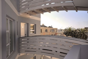 A modern balcony with a white railing, a glass door, and a view of neighboring buildings and palm trees.