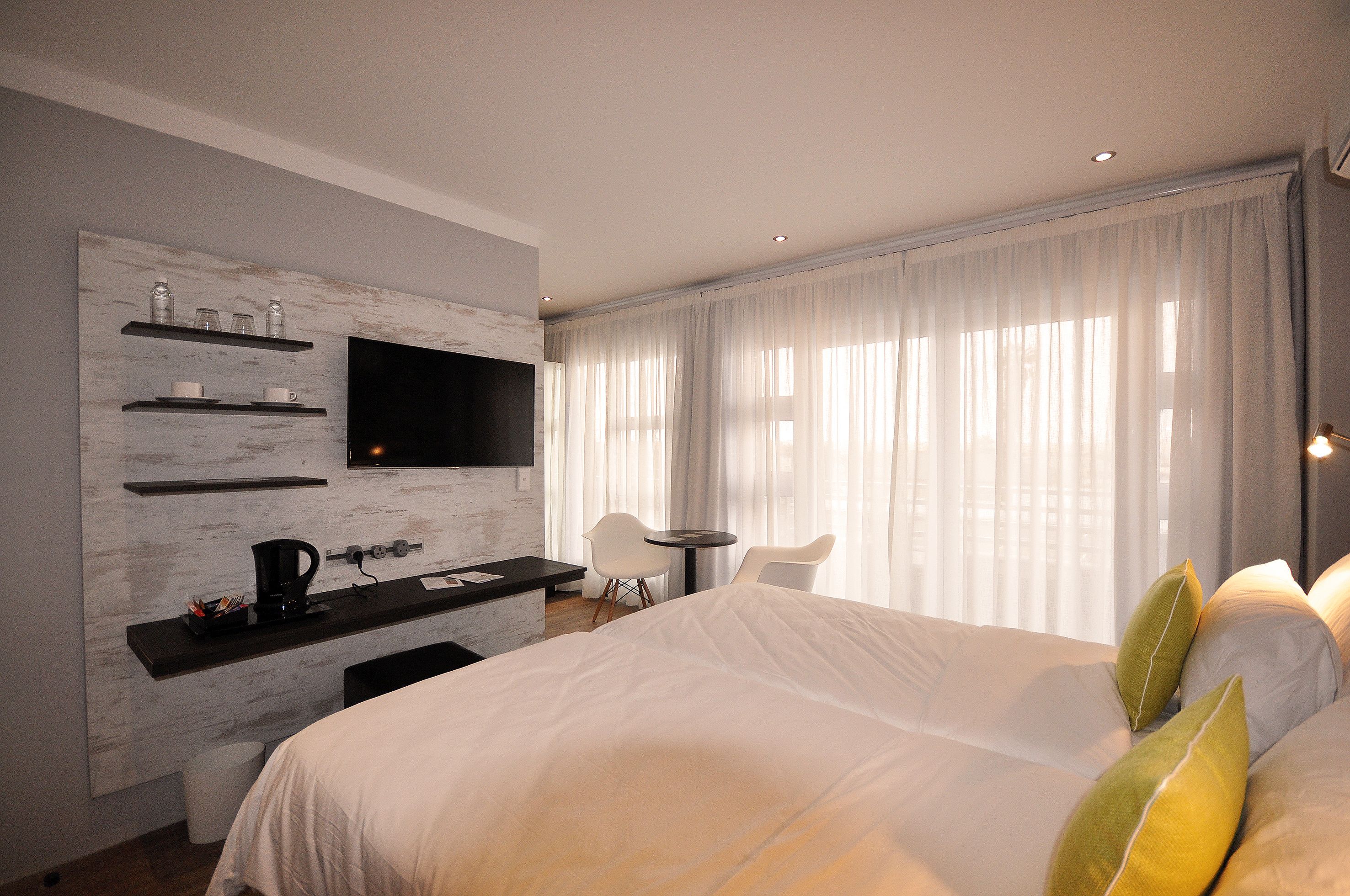 A modern hotel room with a large bed, a flat-screen TV mounted on a wall, a small desk with a chair, and a window with sheer curtains.