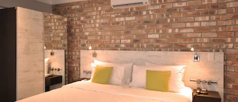 Una habitación de hotel con pared de ladrillo, una cama grande con ropa blanca y dos almohadas, mesitas de noche con lámparas y un equipo de aire acondicionado fijado en la pared.