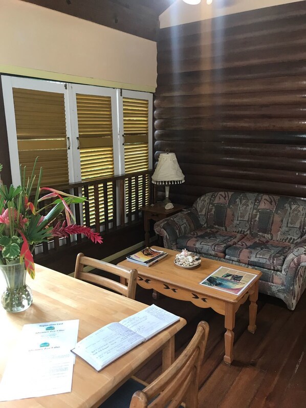 Una sala de estar acogedora con un sofá de madera, una mesa de centro y una mesa de comedor con sillas. Hay una lámpara en una mesita auxiliar y una ventana con persianas.