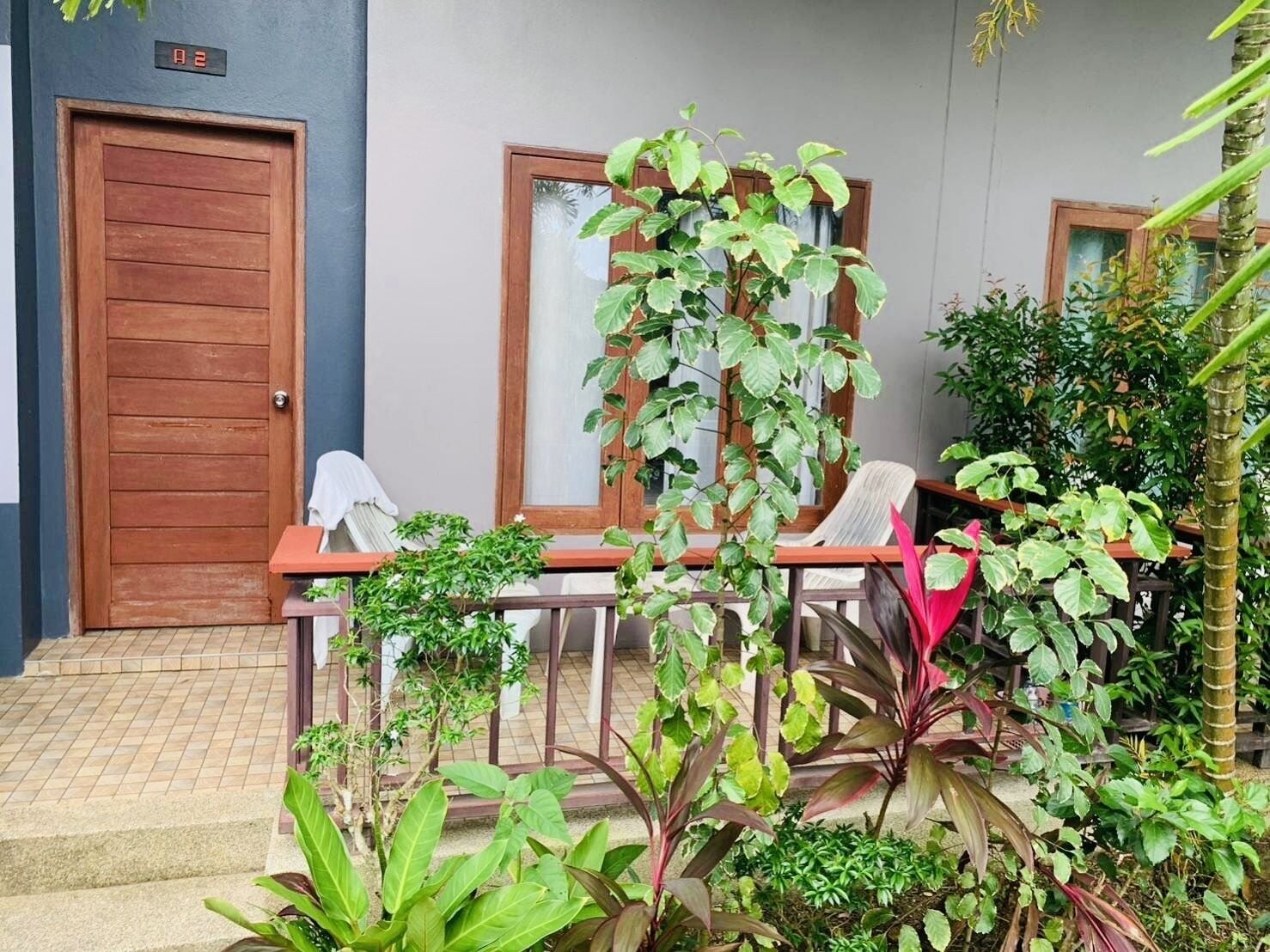 Una puerta de madera con una pantalla digital, un área de estar al aire libre techada con sillas y diversas plantas.