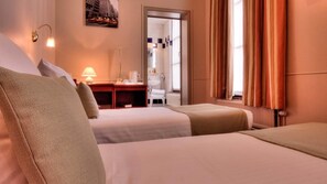 A hotel room with two beds, a desk with a lamp, a mirror, and a view of the city through the window. - Hotel Infotel (Mons)