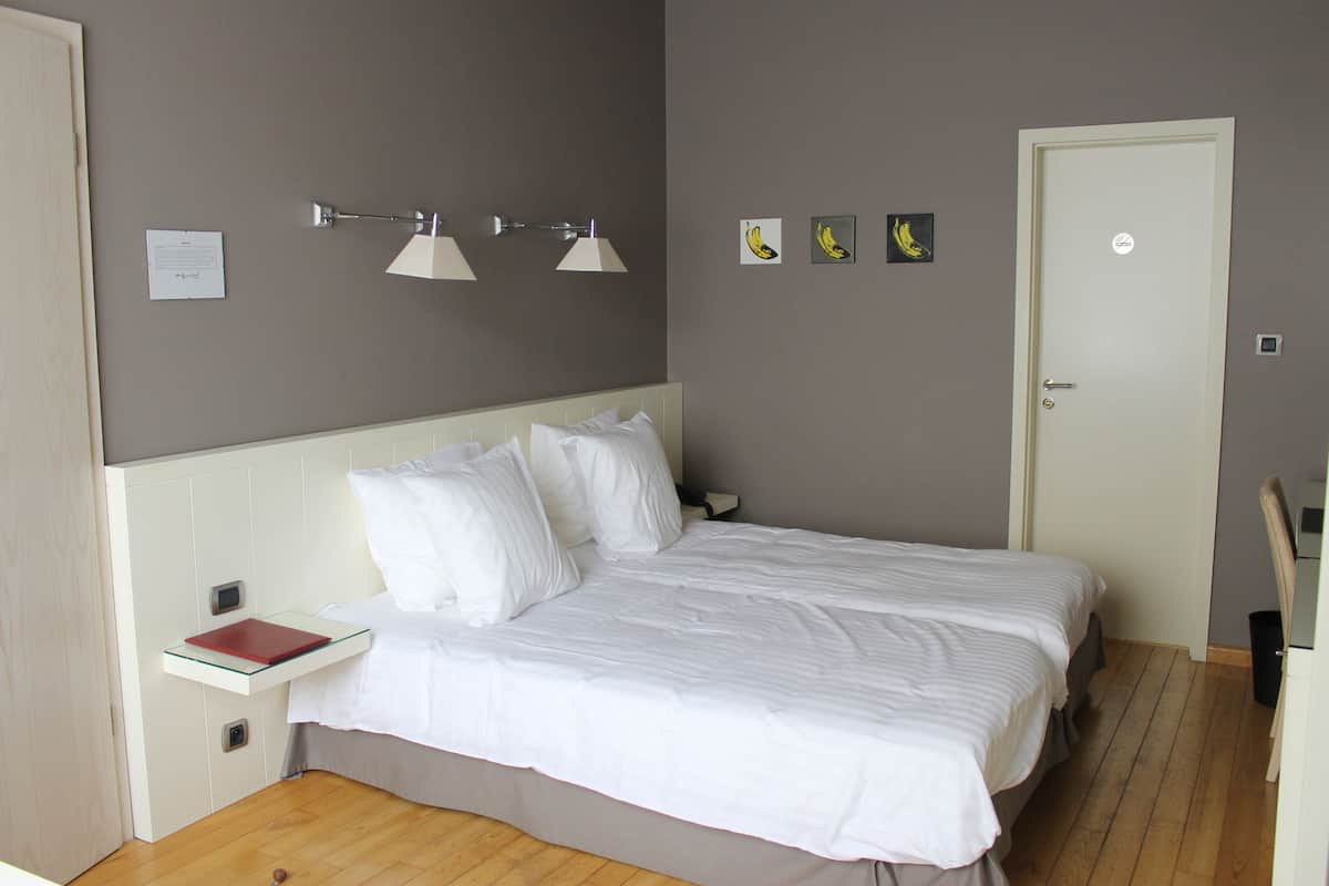 A neatly made bed with white linens, a bedside table with a red book, and wall-mounted lamps.