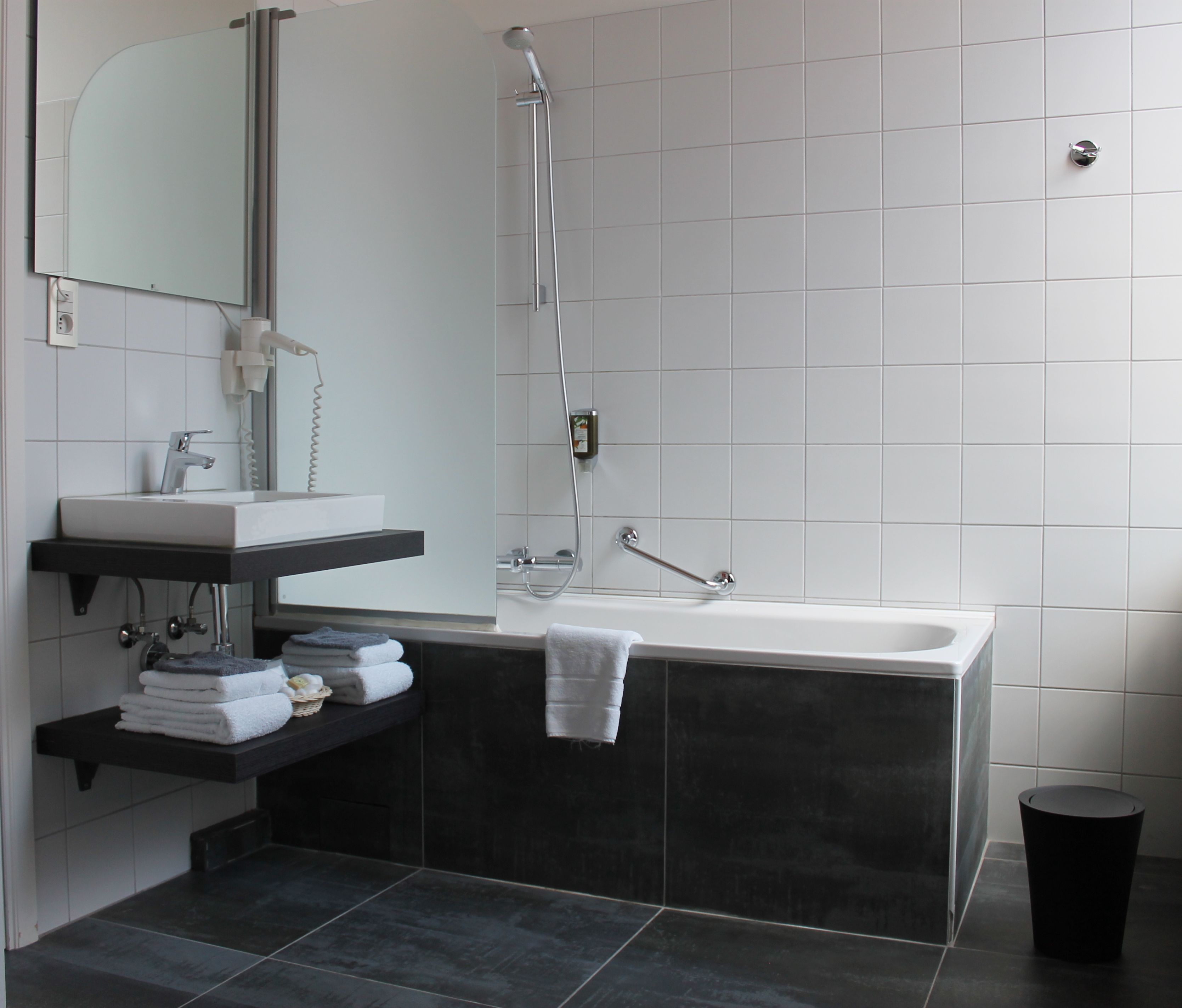 Un baño moderno con un lavamanos blanco, baldosas oscuras, una bañera grande y un espejo sobre el lavamanos.