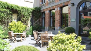 An outdoor patio with wooden tables and chairs, surrounded by greenery and a brick building. - Hotel d'Alcantara (Tournai)