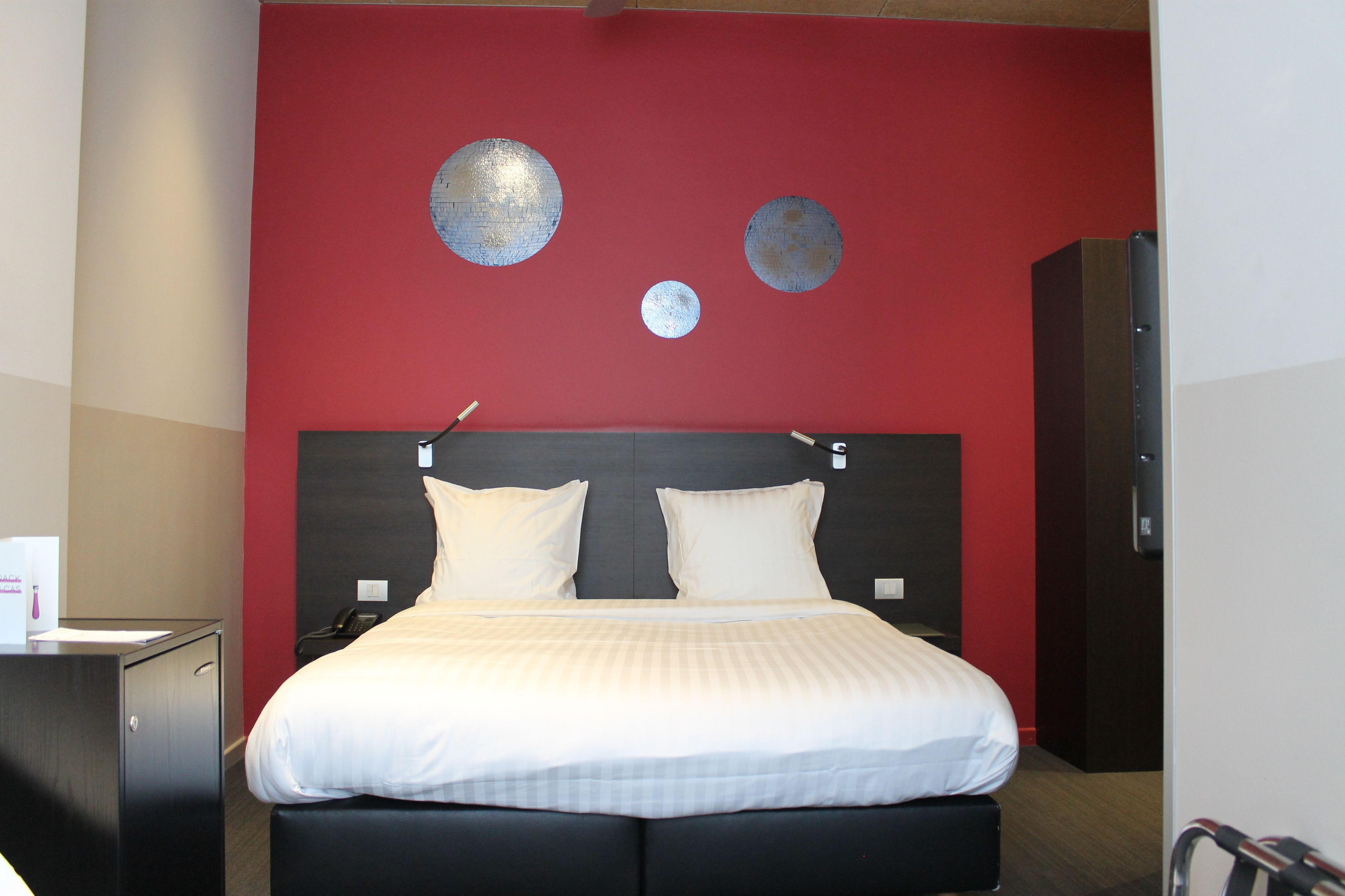Quarto de hotel com uma cama grande, duas almofadas, cabeceira e parede de destaque vermelha com decorações circulares.