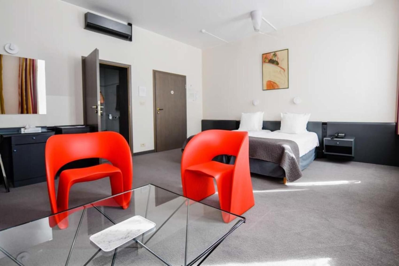 Una camera d'albergo moderna con un tavolino in vetro, due sedie rosse, un letto con biancheria bianca, un quadro appeso e una scrivania con un computer.