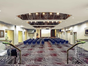 Un salón de conferencias con alfombra estampada, sillas azules, un escenario y un diseño moderno de techo.