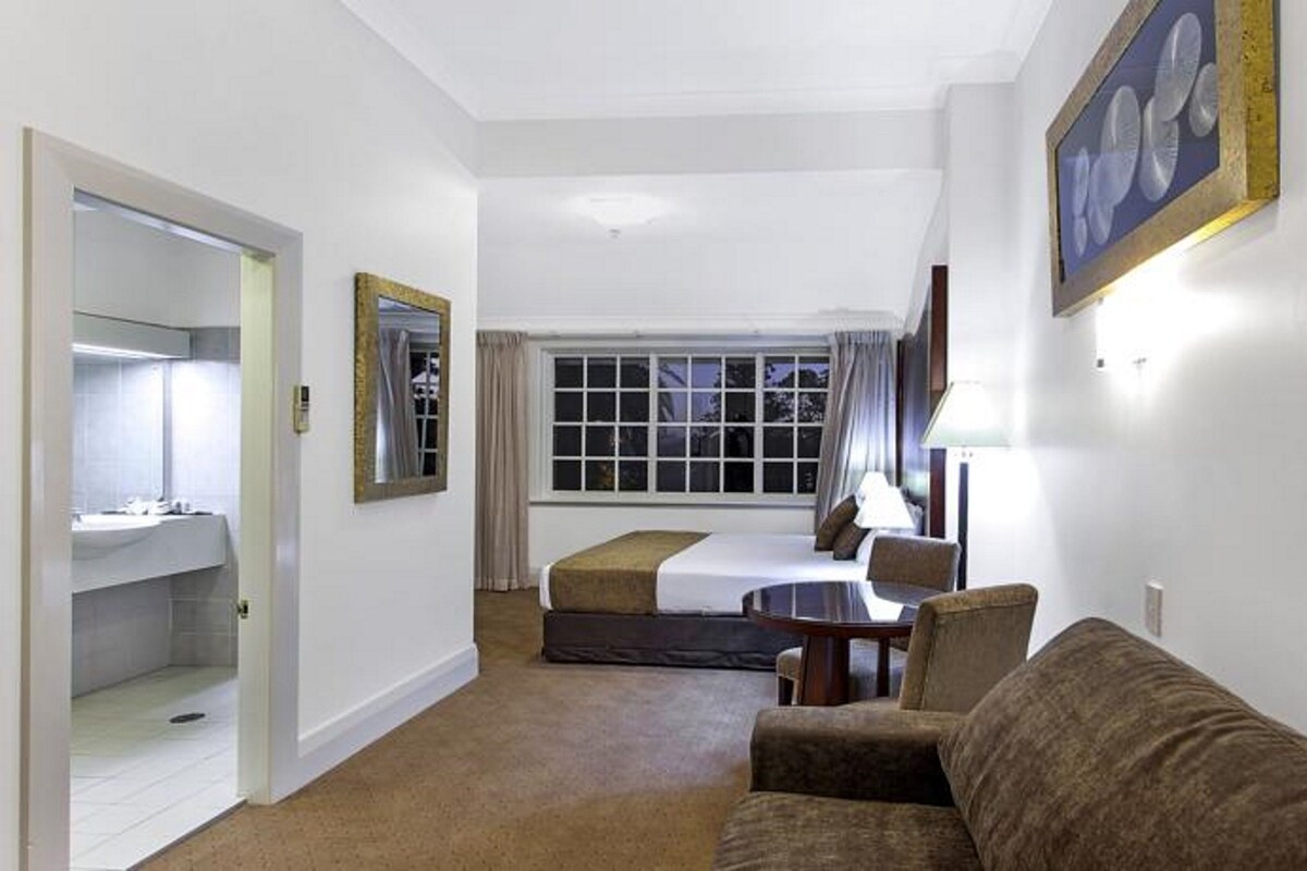 Habitación de hotel con cama, sofá, escritorio, espejo y ventana con cortinas.