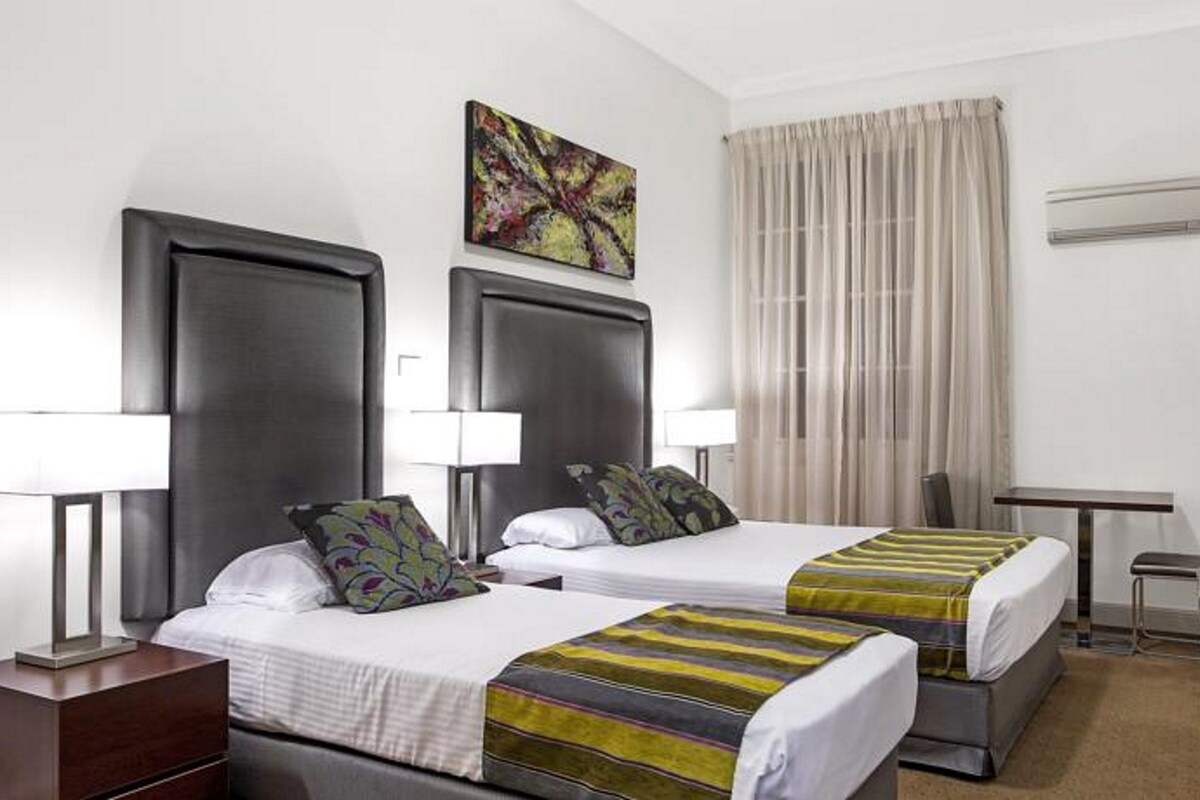Habitación de hotel con dos camas, cada una con cabecera, lámparas de noche y colcha a rayas. Hay un cuadro sobre las camas y una ventana con cortinas.