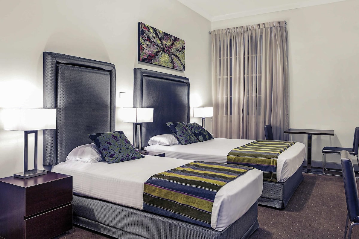 A hotel room with two beds, a desk, a chair, a lamp, and a painting on the wall.
