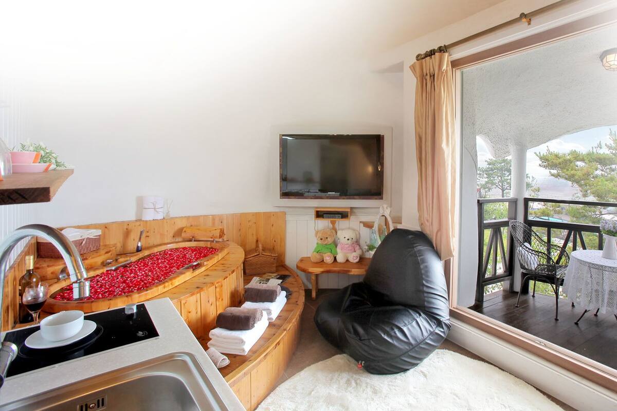 A compact room with a wooden bathtub filled with red flowers, a small table with teddy bears, a TV mounted on the wall, and a balcony with outdoor furniture.