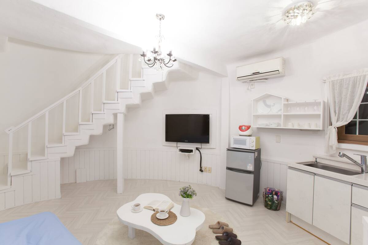A modern, white-themed living room with a staircase, a flat-screen TV, a kitchen area with a refrigerator, and a dining table with a vase of flowers.