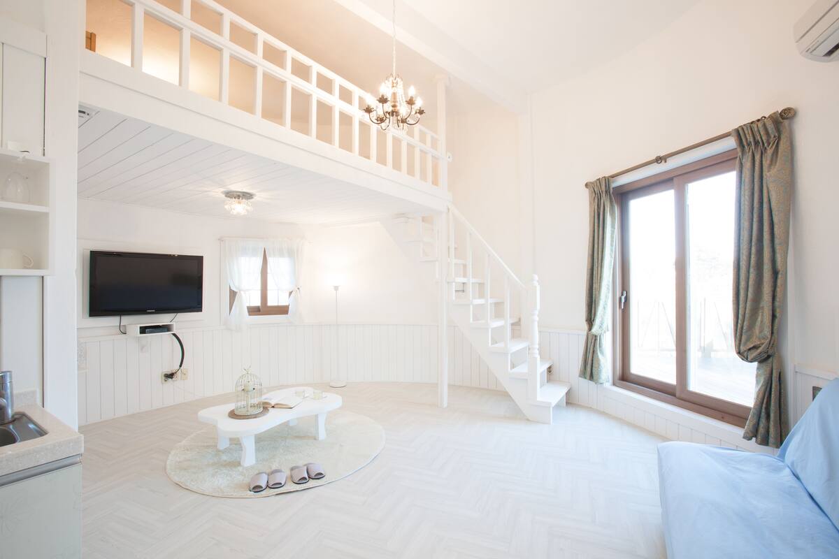 A spacious living area with a white color scheme, a television mounted on the wall, a staircase leading to an upper level, and a large window with curtains.