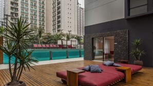 Uma área de estar moderna ao ar livre, com piso de madeira, assentos acolchoados em vermelho e vista para uma piscina e prédios altos ao fundo.
