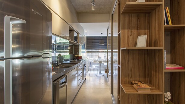 Uma cozinha moderna com eletrodomésticos de aço inoxidável, uma área de jantar com mesa e cadeiras brancas, e prateleiras de madeira com livros e uma pequena planta.