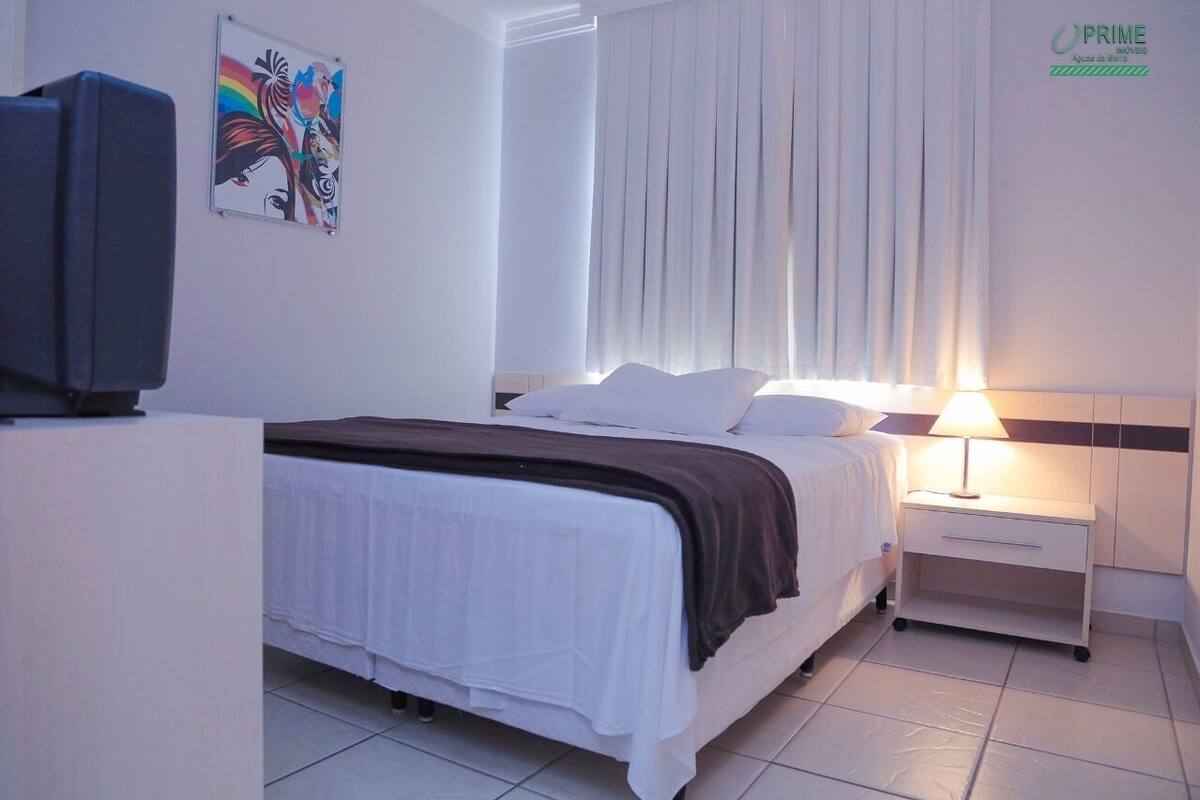 A hotel room with a bed, a nightstand with a lamp, a TV, and a painting on the wall.