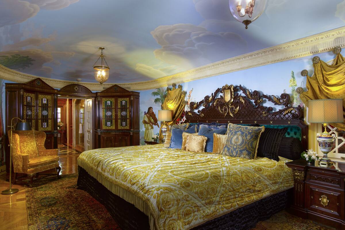 Un dormitorio lujoso con una cama grande, cabecera ornamentada y un mural en el techo.
