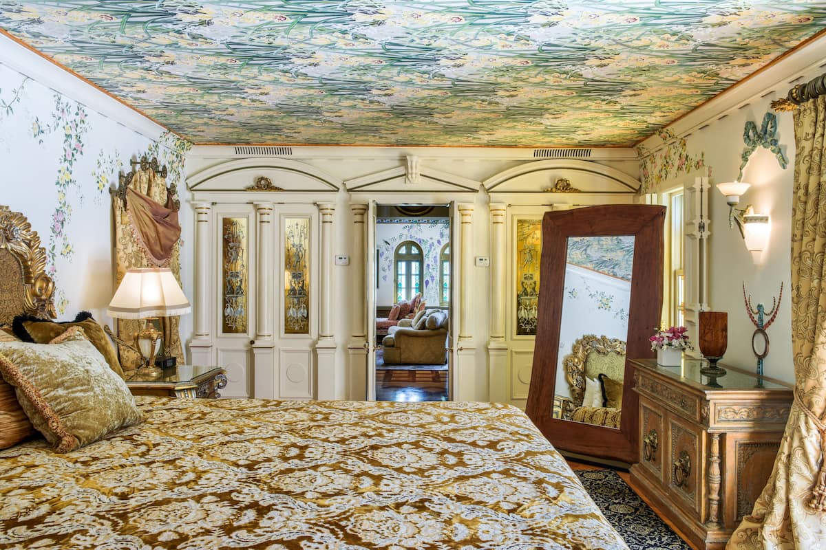 Un dormitorio lujoso con una colcha estampada, muebles ornamentados y un techo decorado con motivos florales.