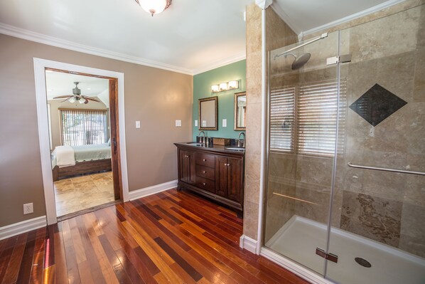 A bathroom with a glass shower enclosure, wooden flooring, a vanity with a mirror, and a door leading to a bedroom. - Chateau Annette (Lexington)