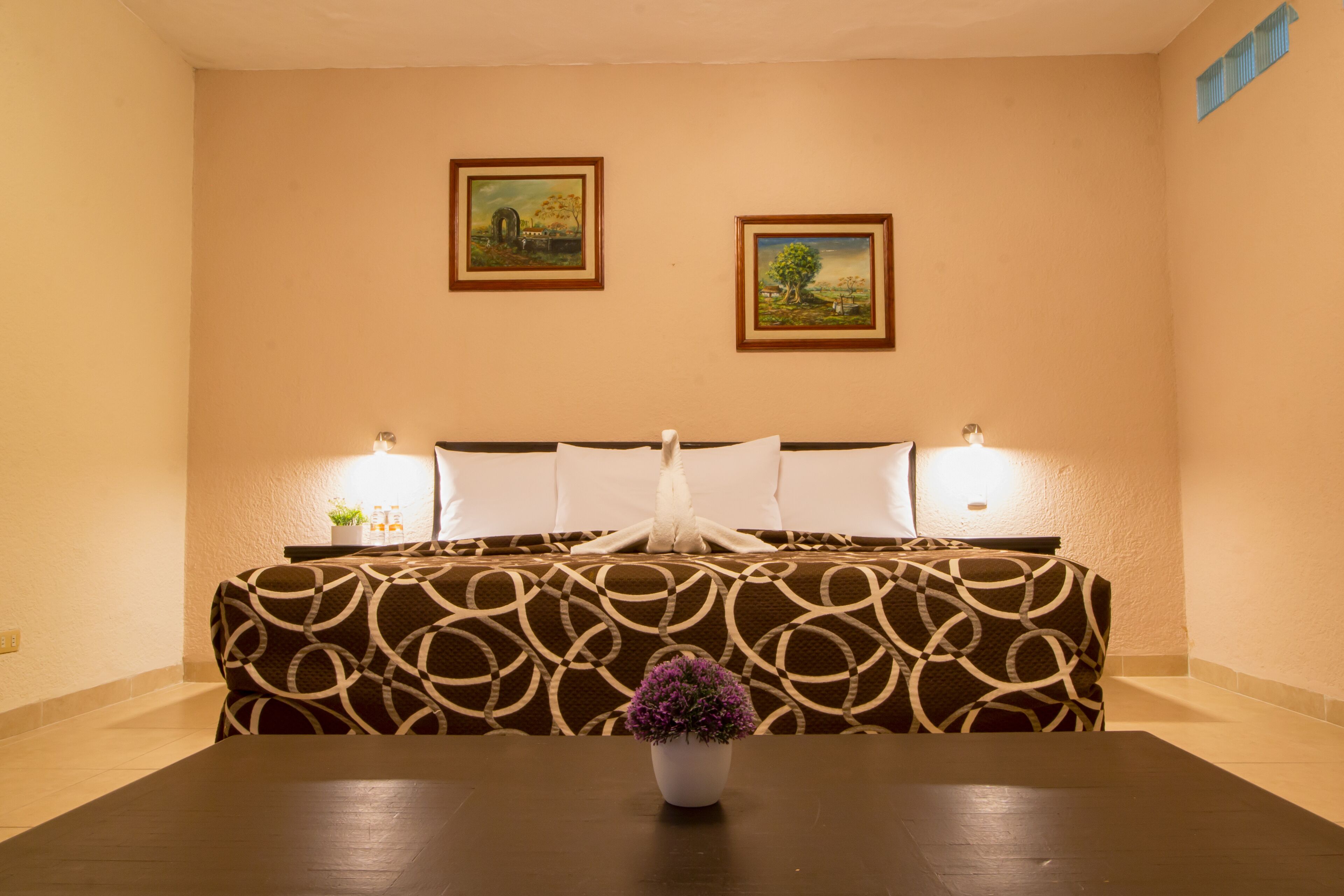 Habitación de hotel con una cama, dos cuadros en la pared, una mesa con un jarrón de flores y una mesita de noche con una lámpara.