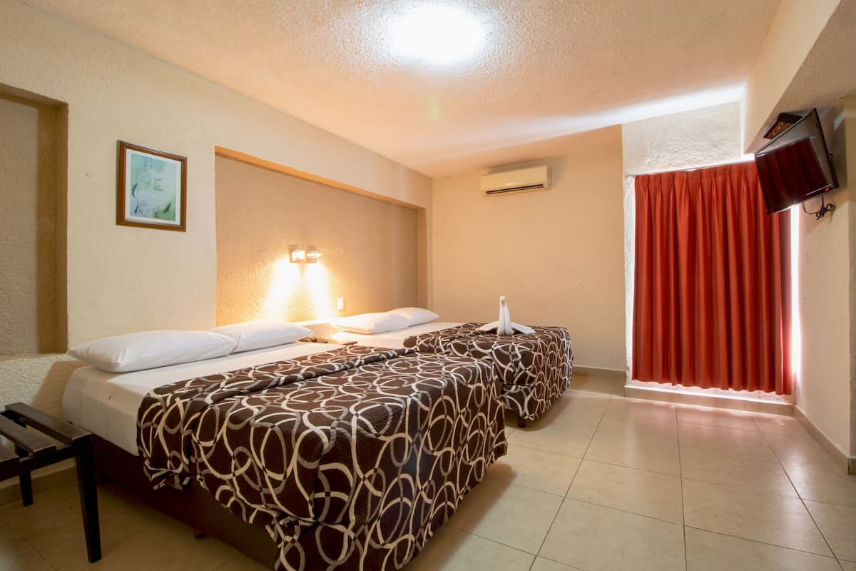 A hotel room with two beds, a bedside table, a lamp, an air conditioning unit, a television, and a window with curtains.