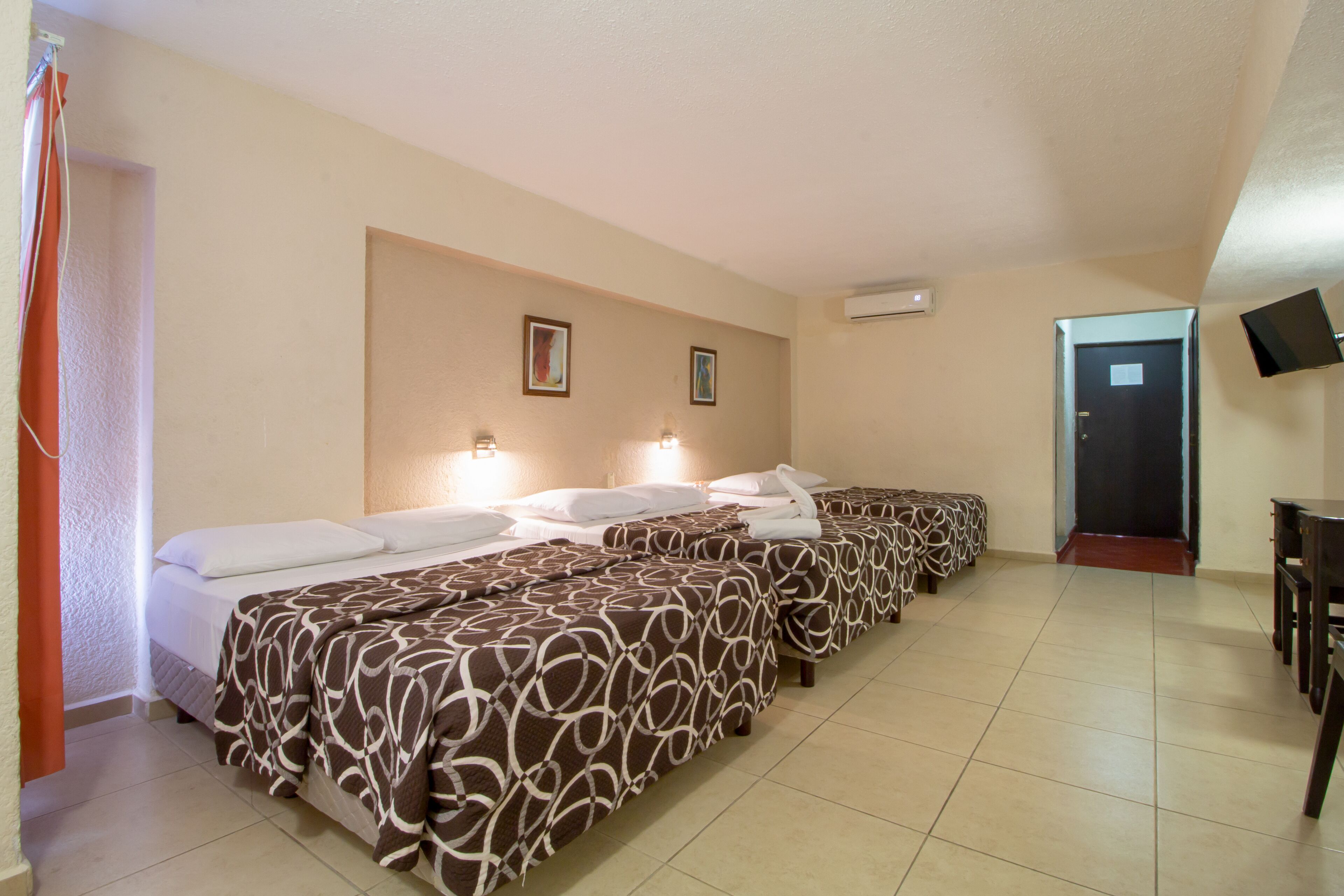 A hotel room with three beds, each with a patterned bedspread, a flat-screen TV mounted on the wall, and a door leading to another room.