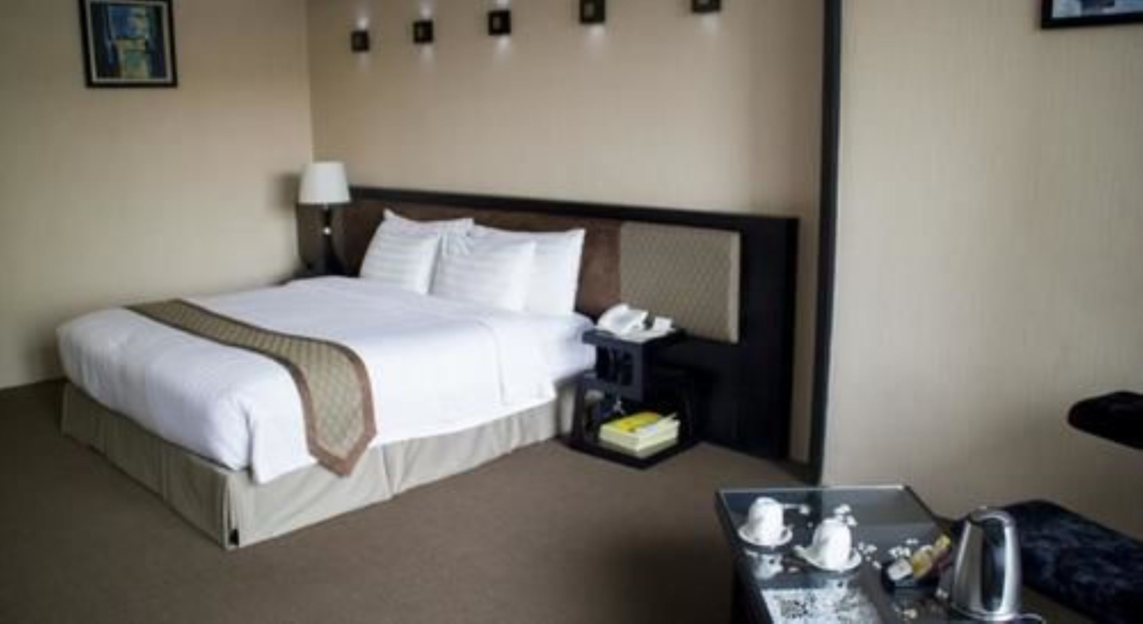 A hotel room with a large bed, a bedside table with a lamp, a telephone, and a small table with a kettle and cups.