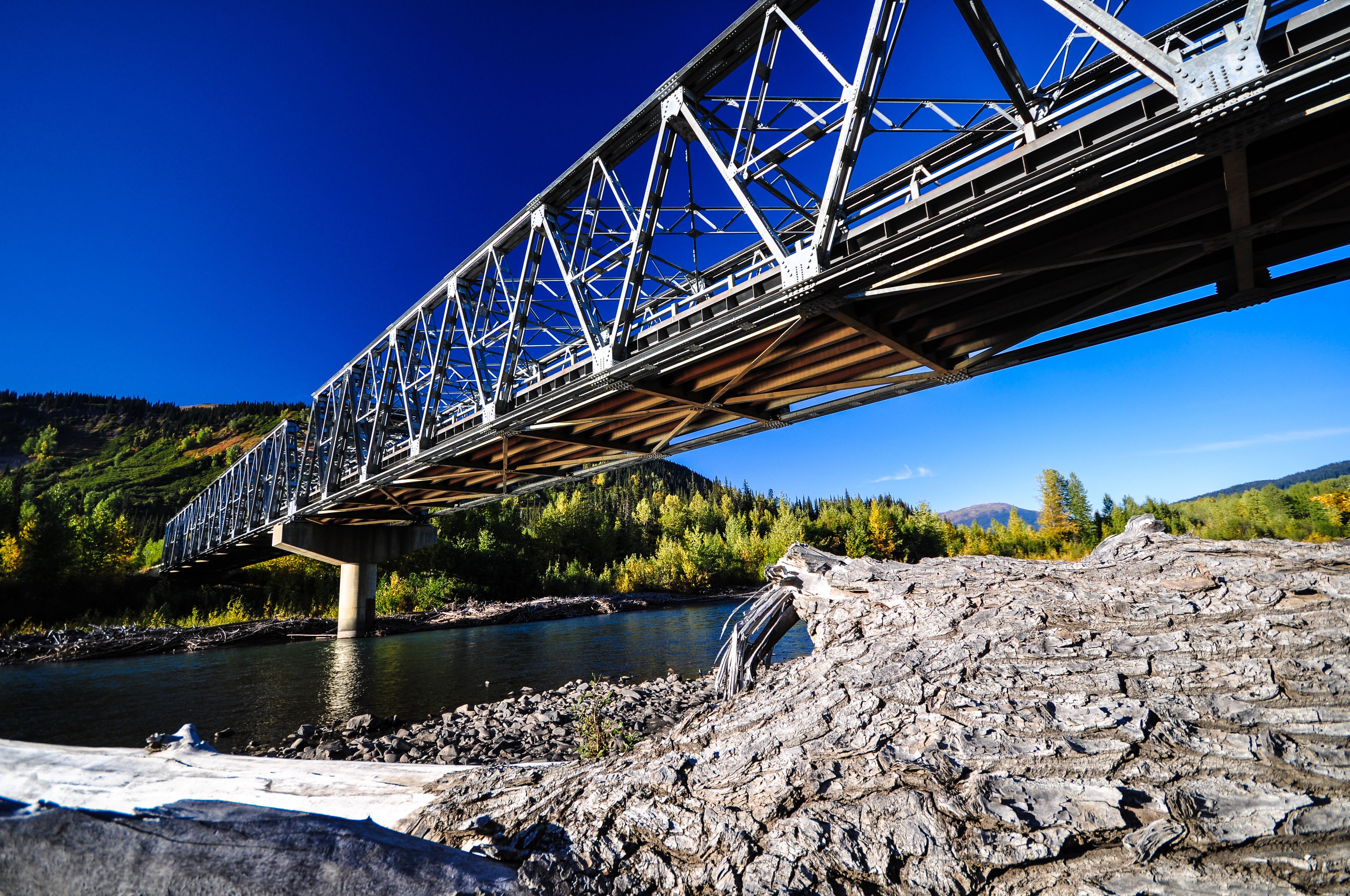 A steel bridge over a river with a rocky foreground and forested hills in the background.