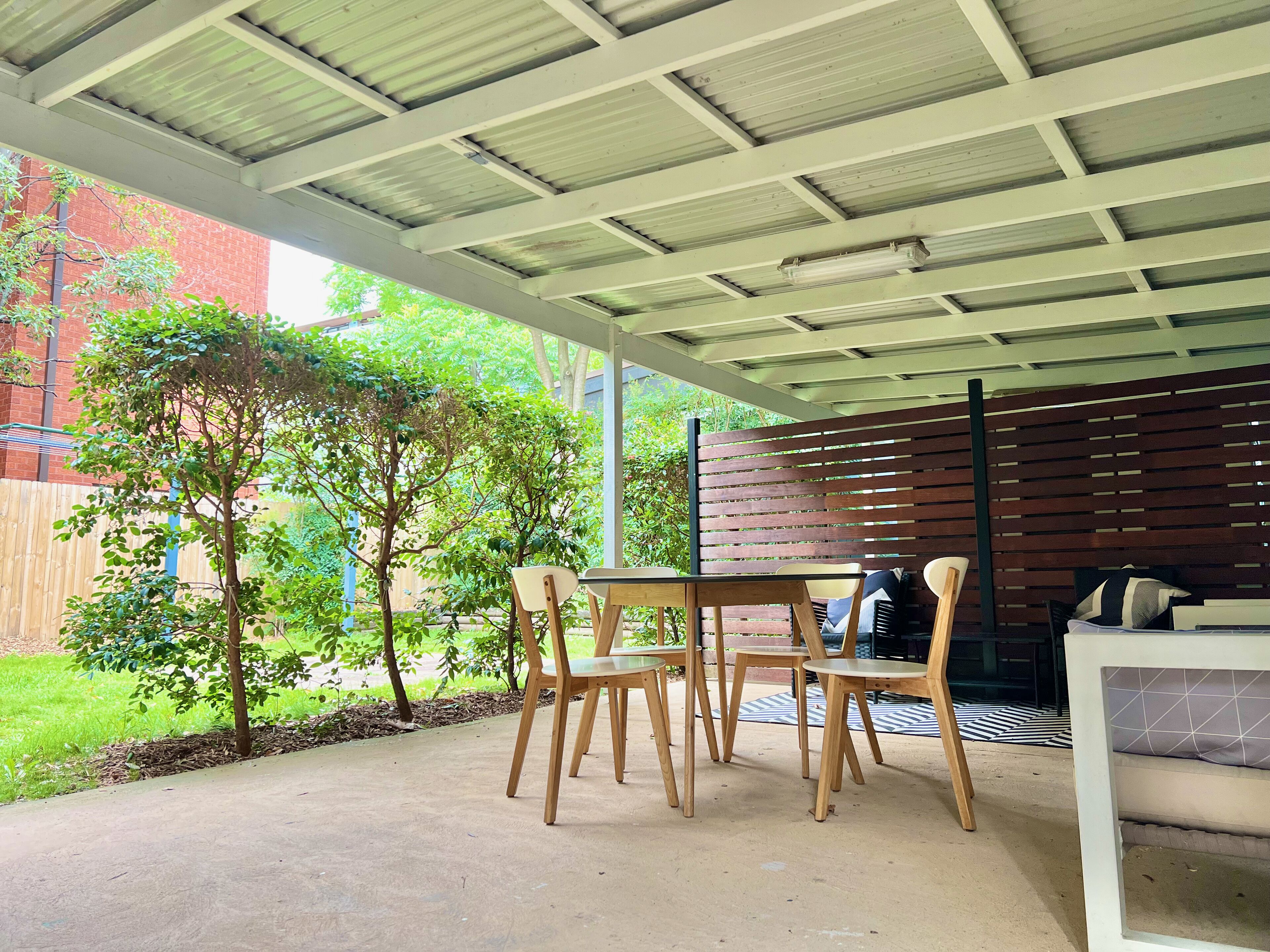 Un'area patio esterna con tettoia, sedie in legno, un tavolo e un divano.