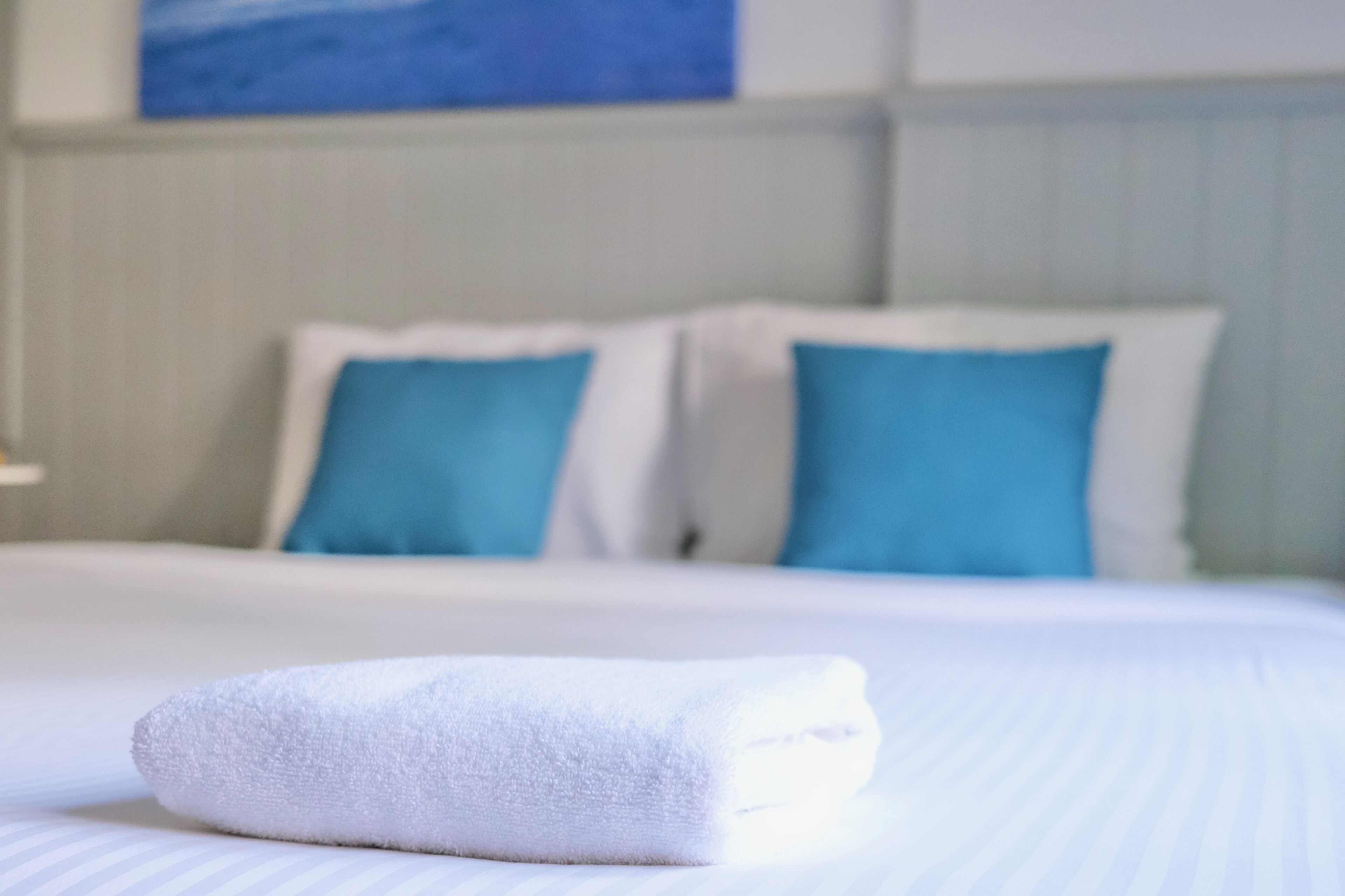 A neatly made bed with white bedding and blue pillows, a white towel on the bed, and a painting on the wall in the background.