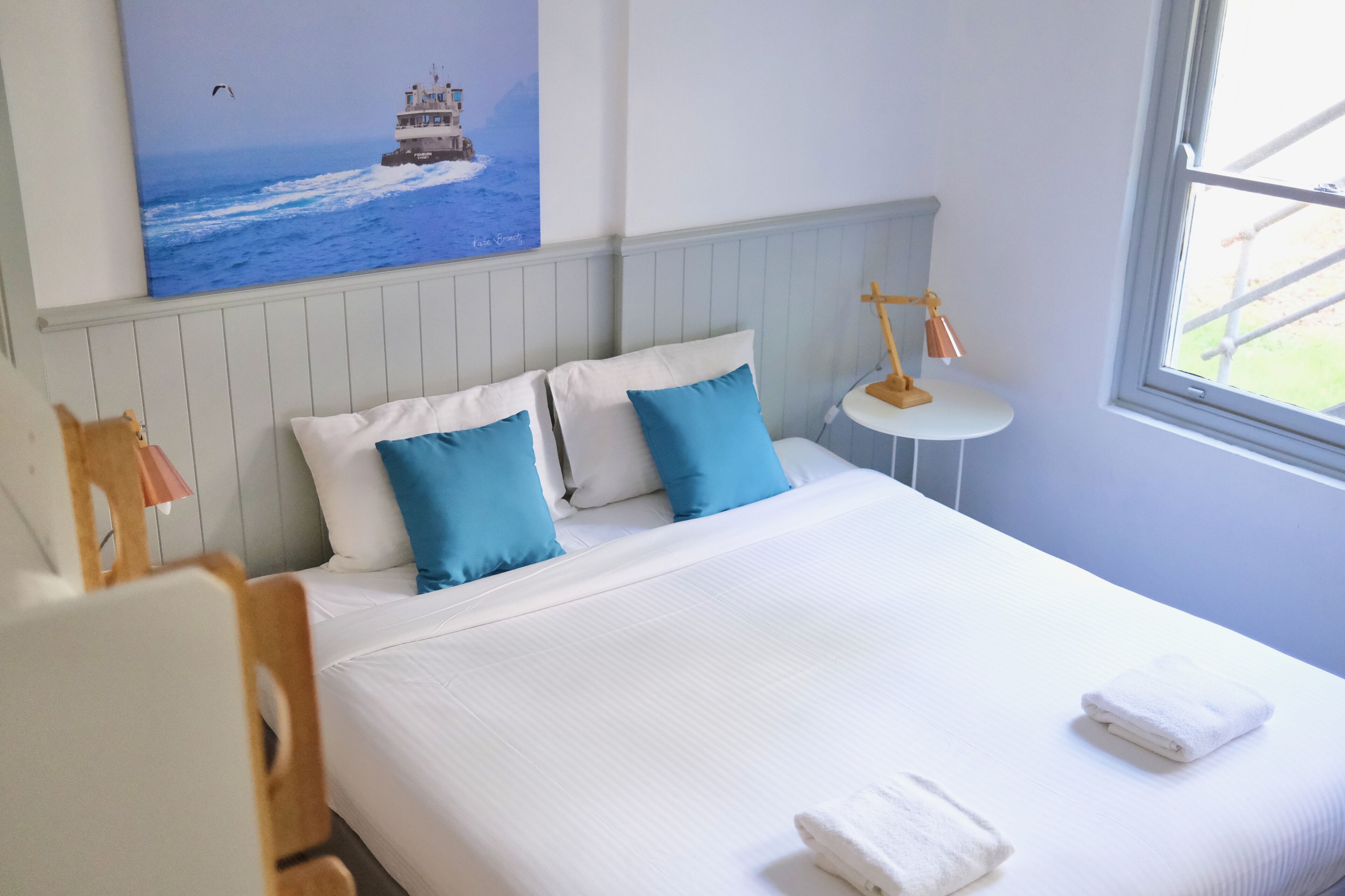 A neatly made bed with white and blue bedding, a small bedside table with a lamp, and a painting of a lighthouse on the wall.