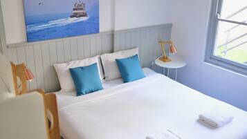 A neatly made bed with white and blue bedding, a small bedside table with a lamp, and a painting of a lighthouse on the wall.