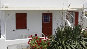 Un edificio blanco con contraventanas rojas y una puerta, rodeado de plantas y un cielo despejado.