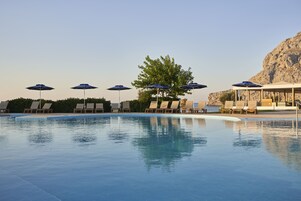 Zona de piscina con sillones de descanso y sombrillas, un árbol y montañas al fondo.