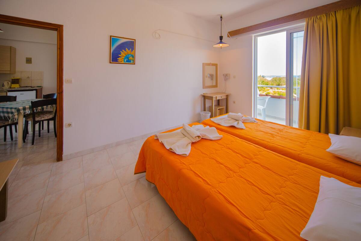 A hotel room with two beds, a small desk, a chair, a painting, and a balcony with yellow curtains.