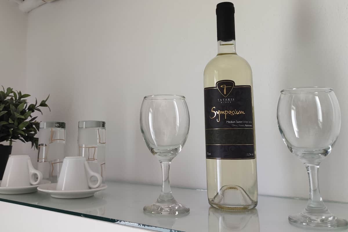 A bottle of Tatakis Wines Symposium Medium Sweet White Wine, two wine glasses, and a set of white teacups with saucers on a glass shelf.