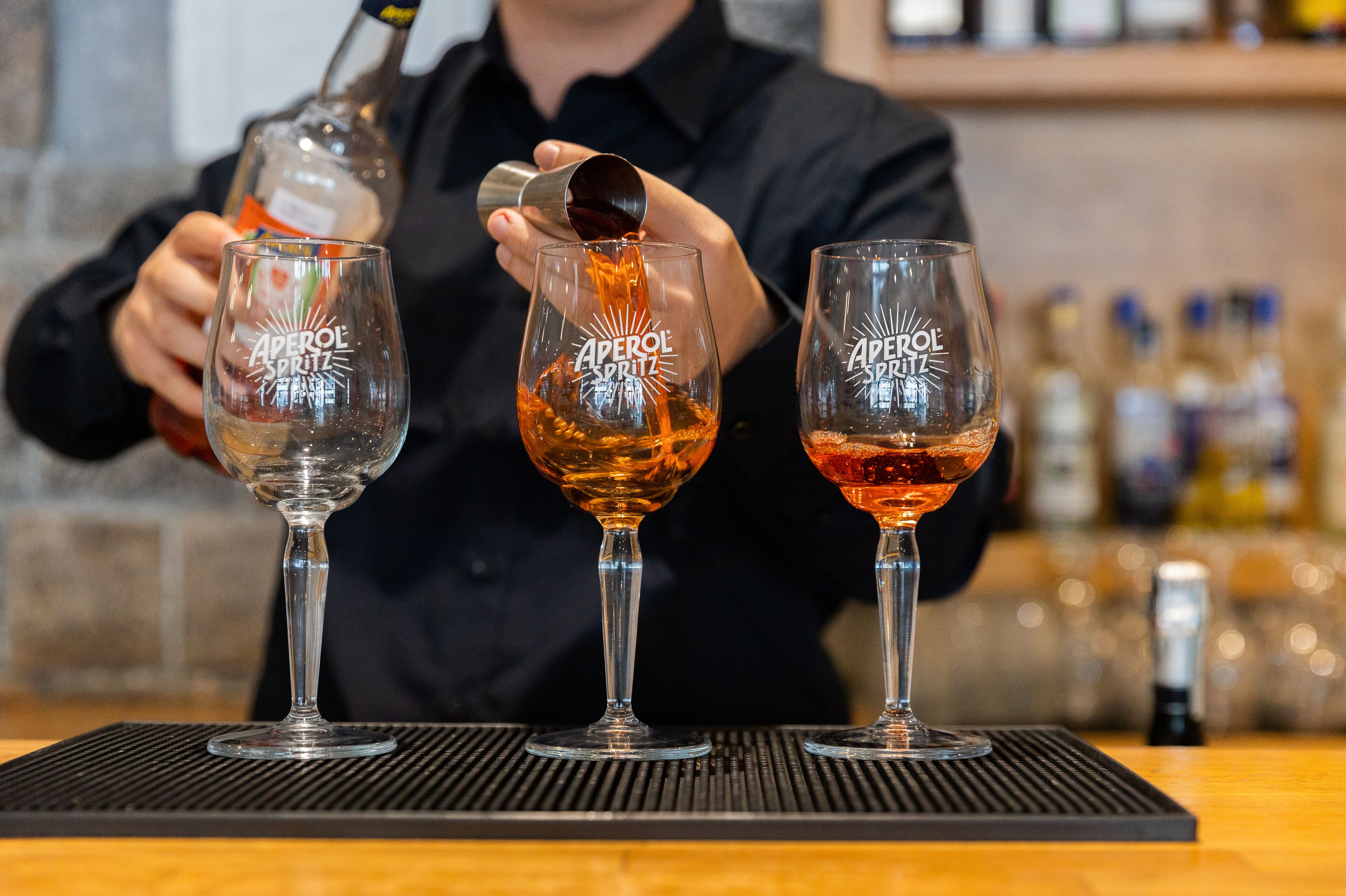 A bartender pouring a drink into a glass labeled "Aperol Spritz" at a bar.
