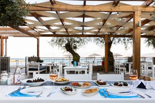An outdoor dining area with a wooden pergola, white tables, and chairs, overlooking a body of water.