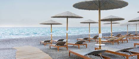 Una playa con sillones de madera, sombrillas de paja y un paseo marítimo de madera, además de una playa de guijarros.