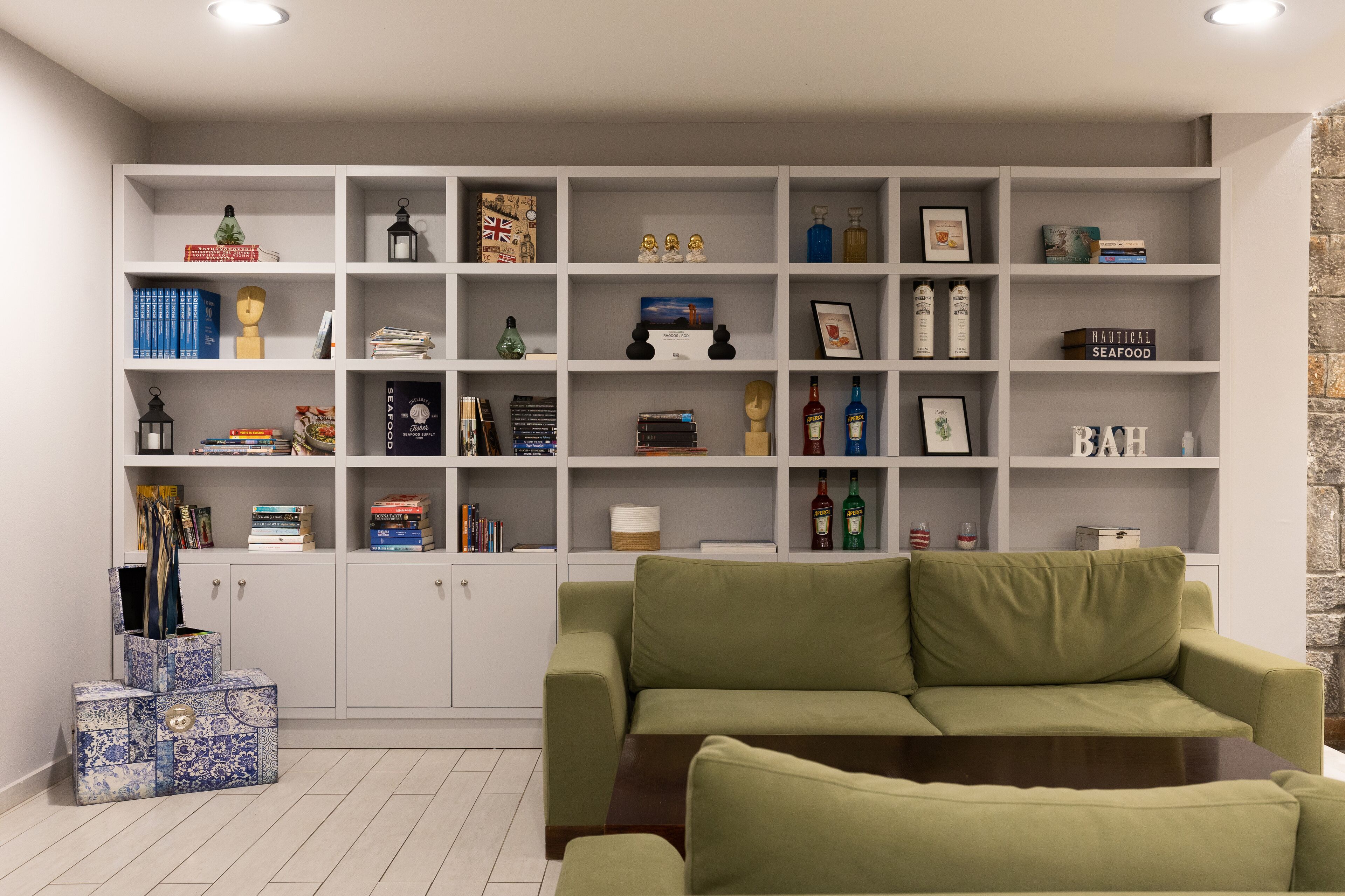 Een woonkamer met een groene bank, een witte boekenkast vol boeken en decoratieve objecten, en een stenen muur.