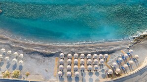 An aerial view of a beach with rows of umbrellas and sun loungers, a rocky shoreline, and clear blue water. - Ariadne Beach - Adults Only (Agios Nikolaos)