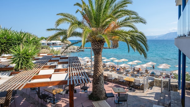 A beachside patio with outdoor seating, palm trees, and a view of the sea. - Ariadne Beach - Adults Only (Agios Nikolaos)