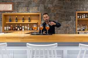 Zona de bar con un bartender preparando bebidas, estantes de madera con botellas y sillas blancas.