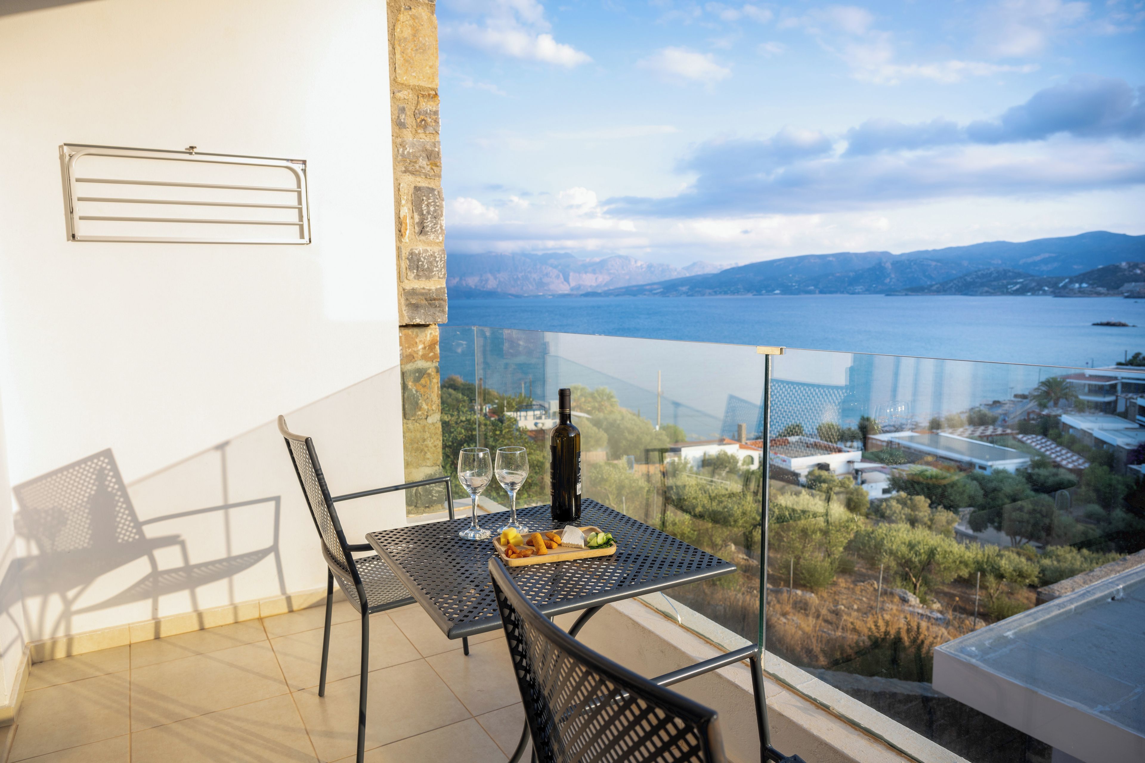 A balcony with a table set for two, a bottle of wine, and a plate of food. The balcony overlooks a body of water and a mountainous landscape.