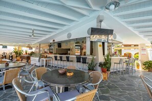 An outdoor cafe area with round tables and chairs, a bar counter, and potted plants. - Kamari Corfu Hotel & Apartments (Corfu)