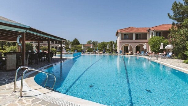 A swimming pool with lounge chairs and umbrellas, surrounded by buildings with red-tiled roofs. - Kamari Apartments (Corfu)